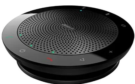 A professional speakerphone rests on a conference table in a modern meeting room.