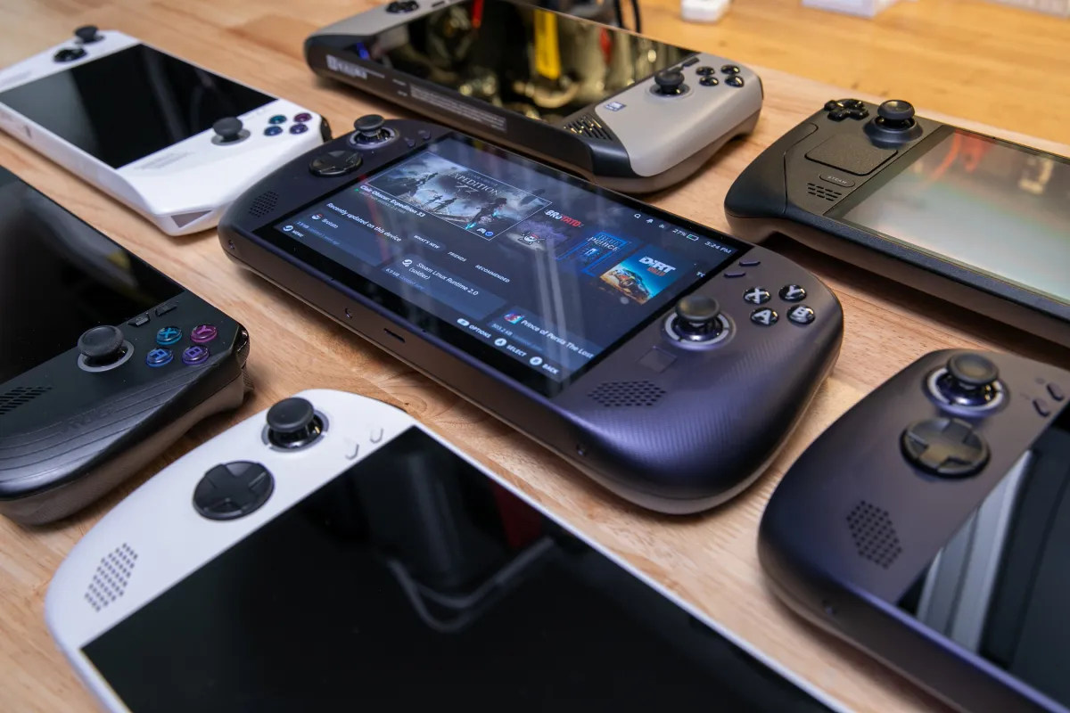 A handheld gaming PC rests on a desk. The screen displays a game interface.