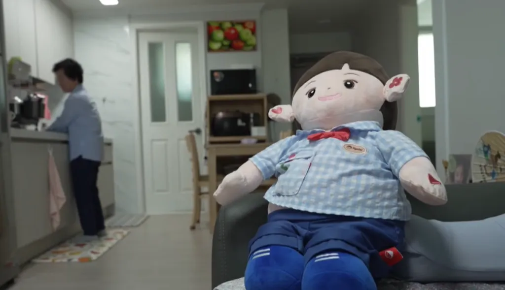 An elderly woman sits beside a childlike AI companion doll in a small apartment.
